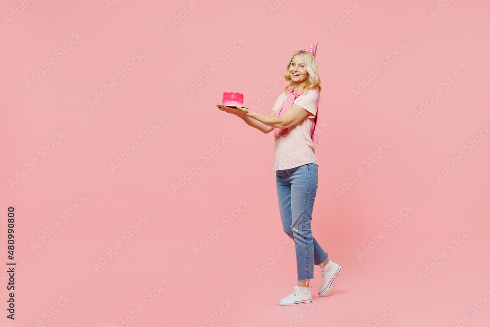 Full body side view elderly happy smiling cheerful satisfied woman 50s wear t-shirt birthday hat hold give cake go isolated on plain pastel pink background studio Celebration of holiday party concept. Full body side view elderly happy smiling cheerful satisfied woman 50s wear t-shirt birthday hat hold give cake go isolated on plain pastel pink background studio Celebration of holiday party concept.