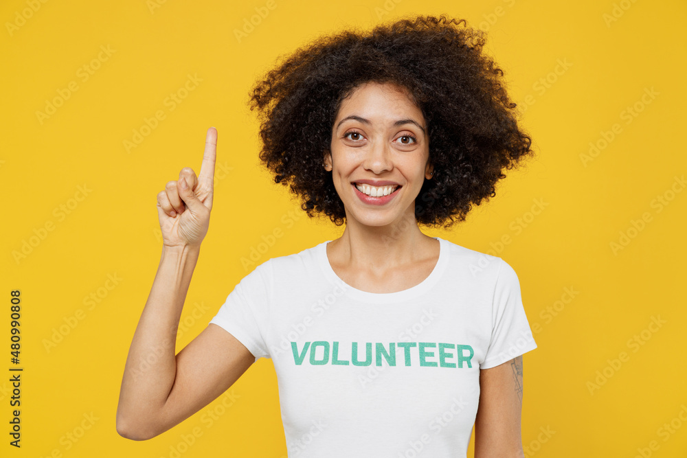 Young woman of African American ethnicity in white volunteer t-shirt holding index finger up with great new idea isolated on plain yellow background. Voluntary free work assistance help grace concept. Young woman of African American ethnicity in white volunteer t-shirt holding index finger up with great new idea isolated on plain yellow background. Voluntary free work assistance help grace concept.