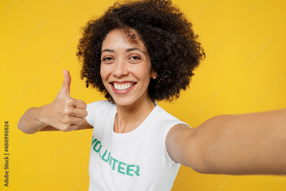 Close up young woman of African American ethnicity wears white volunteer t-shirt doing selfie shot pov on mobile phone show thumb up isolated on plain yellow background. Voluntary free help concept Close up young woman of African American ethnicity wears white volunteer t-shirt doing selfie shot pov on mobile phone show thumb up isolated on plain yellow background. Voluntary free help concept