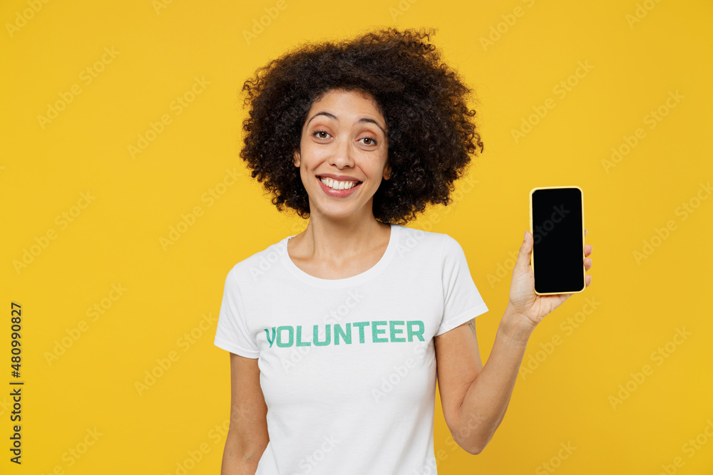 Young woman of African American ethnicity wear white volunteer t-shirt hold use mobile cell phone with blank screen workspace area isolated on plain yellow background Voluntary free work help concept Young woman of African American ethnicity wear white volunteer t-shirt hold use mobile cell phone with blank screen workspace area isolated on plain yellow background Voluntary free work help concept