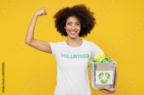 Young fun woman of African American ethnicity wear white volunteer t-shirt hold waste sorting boxes show muscles isolated on plain yellow background. Voluntary free work assistance help grace concept