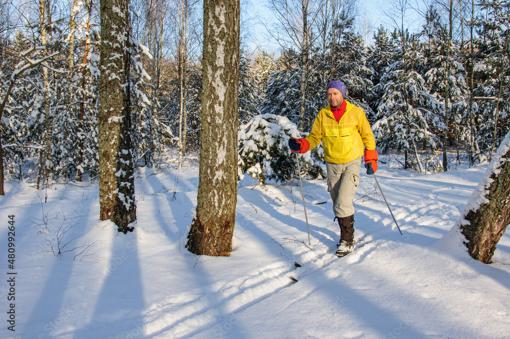 A man in a yellow jacket is walking on snowy woods on wooden skis