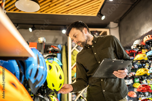 A male bike shop manager makes an inventory of sports helmets in a bike shop. The owner of a sports store with a clipboard in his hands checks the prices of bicycle helmets in the showcase