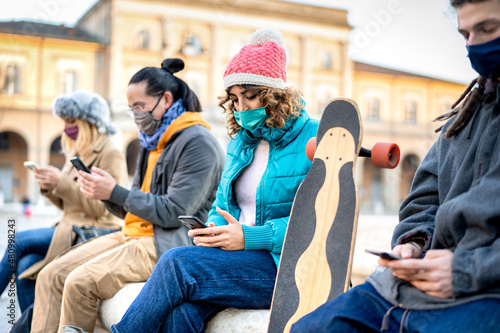 Milenial friends with face mask watching news on smart phone about omicron emergency - Worried guys and girls looking updates on mobile smartphone - College students seating at city center main square photo