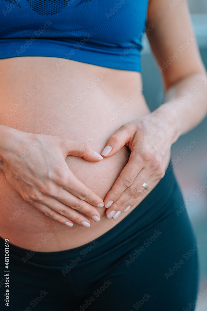 Close up of pregnant woman in blue sports bra and black leggings holding hands on her naked belly making heart symbol with her hands. Love, care, Valentines Day Close up of pregnant woman in blue sports bra and black leggings holding hands on her naked belly making heart symbol with her hands. Love, care, Valentines Day