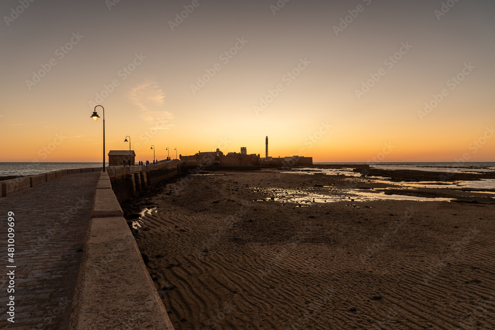 Pedestrian promenade to an islet with the lighthouse and San Sebastian Castle in the old town near La Caleta beach at sunset, Cadiz, Andalusia, Spain