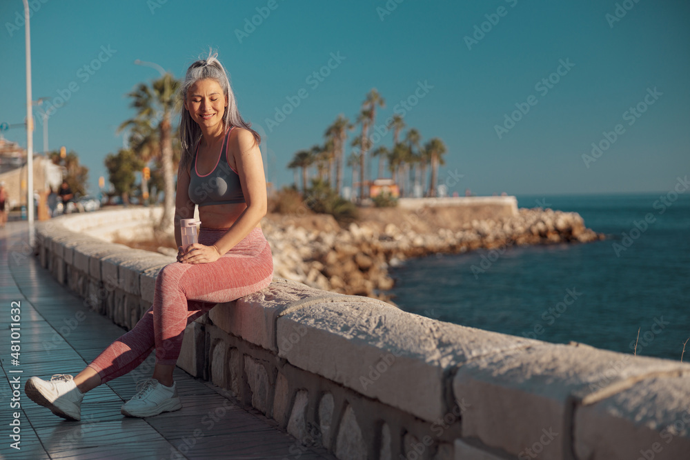 Smiling sporty lady in sports top and pink leggings sitting by the seashore and holding bottle of water