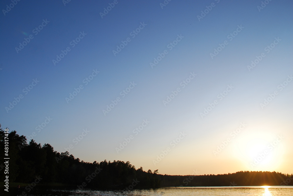 Obraz premium Summer sunset on the shore of a forest lake. The water surface is calm with fine ripples, the sun sets behind the forest on the other side of the lake. The sun's rays are reflected in the water.