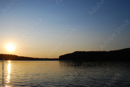 Beautiful sunset on the shore of a forest lake. The sun went down low over the forest growing on the shore of the lake. The surface of the water is almost smooth, the sun's rays are reflected from it.