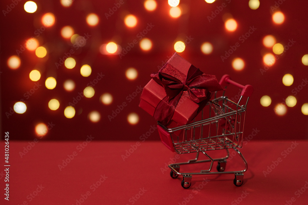 A small grocery cart with a gift box on a red background with out of focus lights in the background. Give gifts with love on Valentine's Day, Christmas and birthdays. Online shopping. Holiday sales