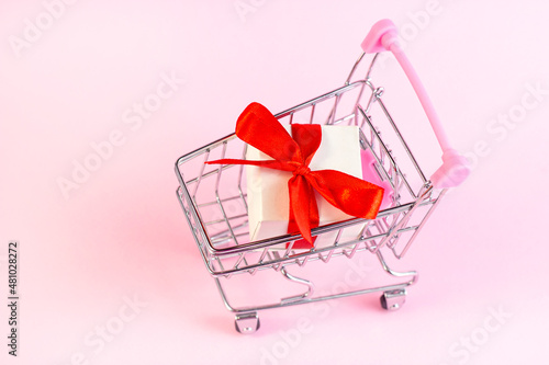 Bright pink shopping trolley push cart with white gift box on light rose background. Valentines day celebration sale, romantic present discounts concept.