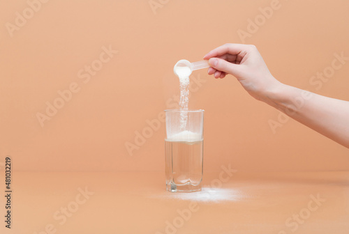Woman pours collagen powder or protein in a glass of water on a beige background. A healthy and anti aging supplement.