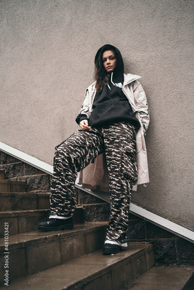 Naklejka premium An upset girl stands against a wall on the street. A girl in a black hoodie. Space for text. A teenager in a black hood is offended.