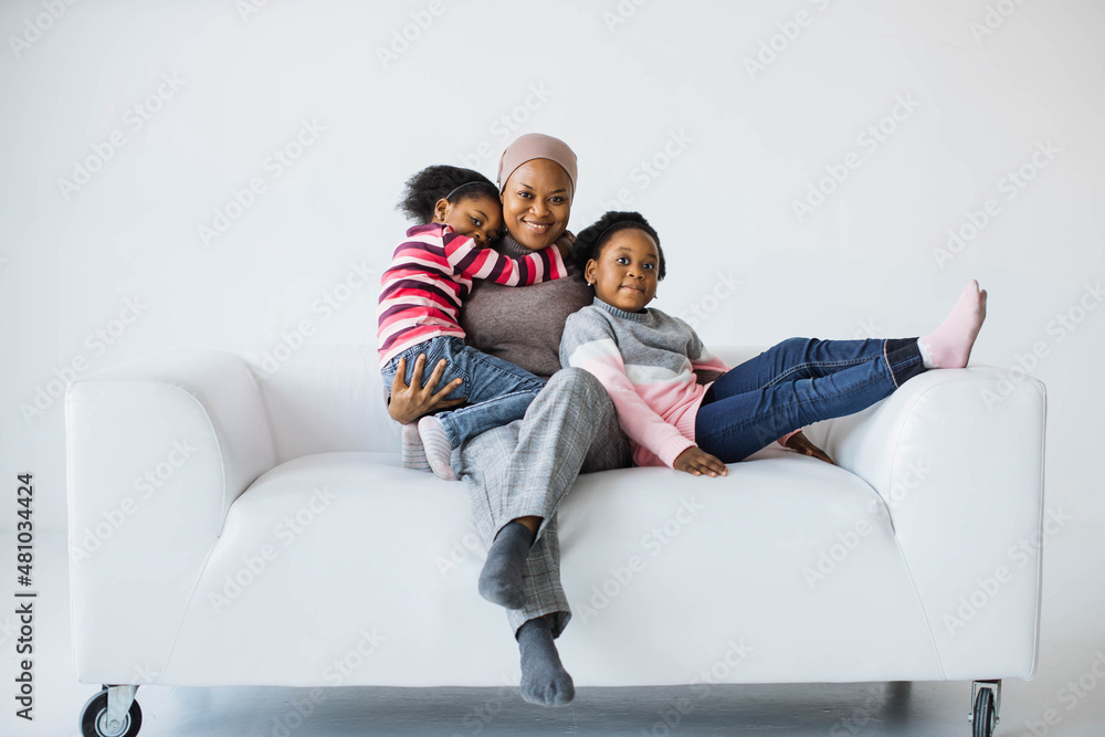 Happy african american muslim mother in headscarf and her two little daughters dressed in casual clothes resting together on comfy couch. White studio background. Family concept. Happy african american muslim mother in headscarf and her two little daughters dressed in casual clothes resting together on comfy couch. White studio background. Family concept.