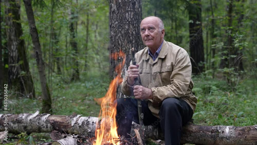 A pensive old traveler is sitting alone in the forest next to a campfire and warming himself A pensive old traveler is sitting alone in the forest next to a campfire and warming himself