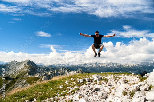 A young man jumps high on top of Vogel mountain in Slovenia. He has the high mountains of the Julian Alps behind him.