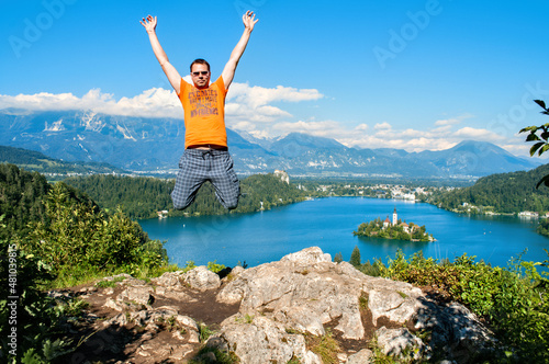 A young man jumps high above Lake Bled in Slovenia. He has the high mountains of the Julian Alps behind him.