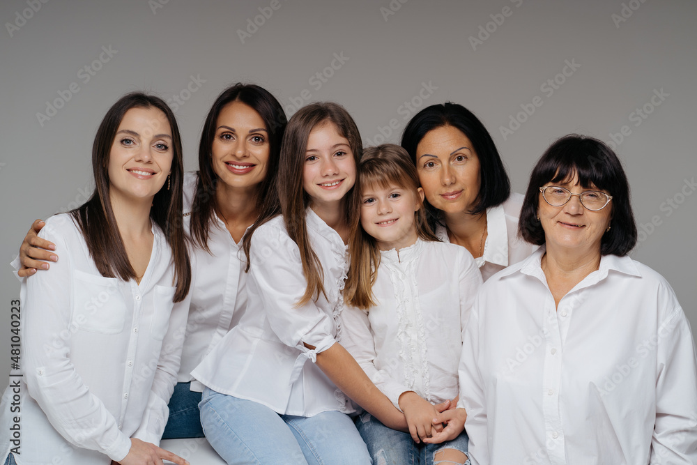 Delighted cute young girls, their moms and grandmother smiling, hugging while standing on gray background wearing jeans and white shirts. Lovely women group Delighted cute young girls, their moms and grandmother smiling, hugging while standing on gray background wearing jeans and white shirts. Lovely women group