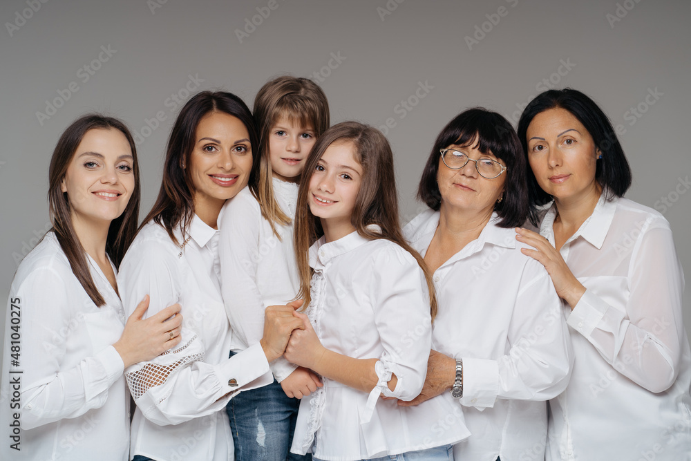 Delighted cute young girls, their moms and grandmother smiling, hugging while standing on gray background wearing jeans and white shirts. Lovely women group Delighted cute young girls, their moms and grandmother smiling, hugging while standing on gray background wearing jeans and white shirts. Lovely women group