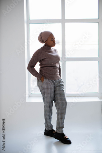 Attractive african american woman in headscarf and casual clothes standing in studio with white background and looking at window. Concept of people and lifestyles.