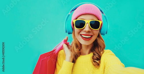 Colorful portrait of stylish happy laughing woman stretching her hand for taking selfie in headphones with shopping bags wearing yellow knitted sweater, pink hat on blue background