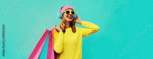 Colorful portrait of stylish smiling young woman listening to music in headphones with shopping bags posing wearing a yellow knitted sweater, pink hat on blue background © rohappy