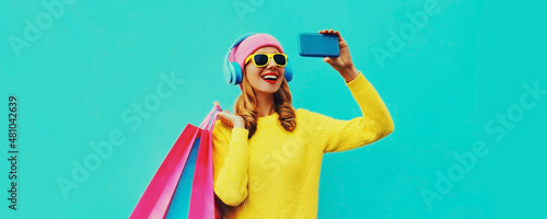 Stylish happy smiling woman taking a selfie by phone in headphones with shopping bags wearing a yellow knitted sweater, pink hat on blue background