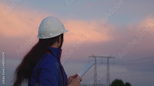 Civil engineer, woman specializing in power supply, works outdoors. Concept of green energy, ecology. Modern technologies. Power engineer in a protective helmet checks the power line, digital tablet