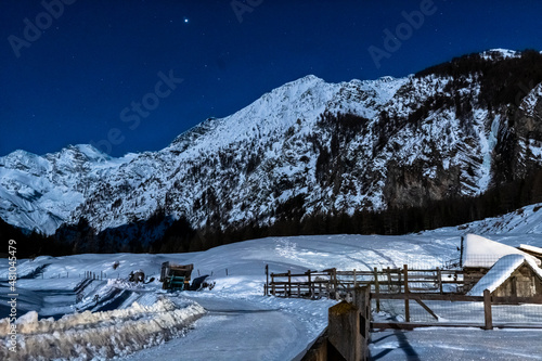 Cogne, Aosta Valley, snowy path on a full moon night with starry sky and mountains in the background