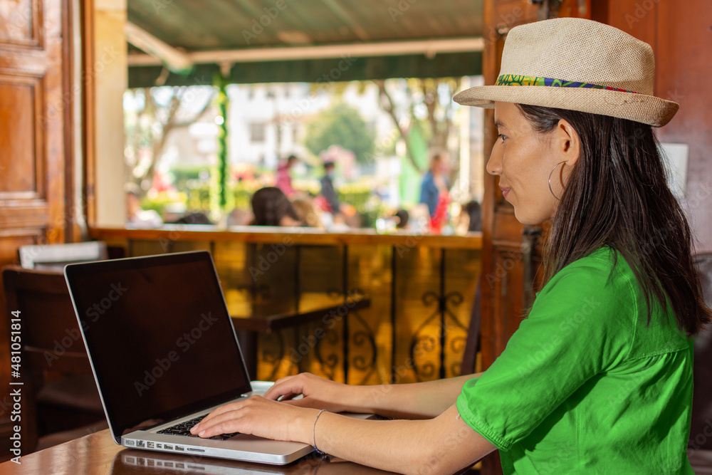 Latin woman wearing a hat and green blouse, sitting in a old coffee shop working on her laptop