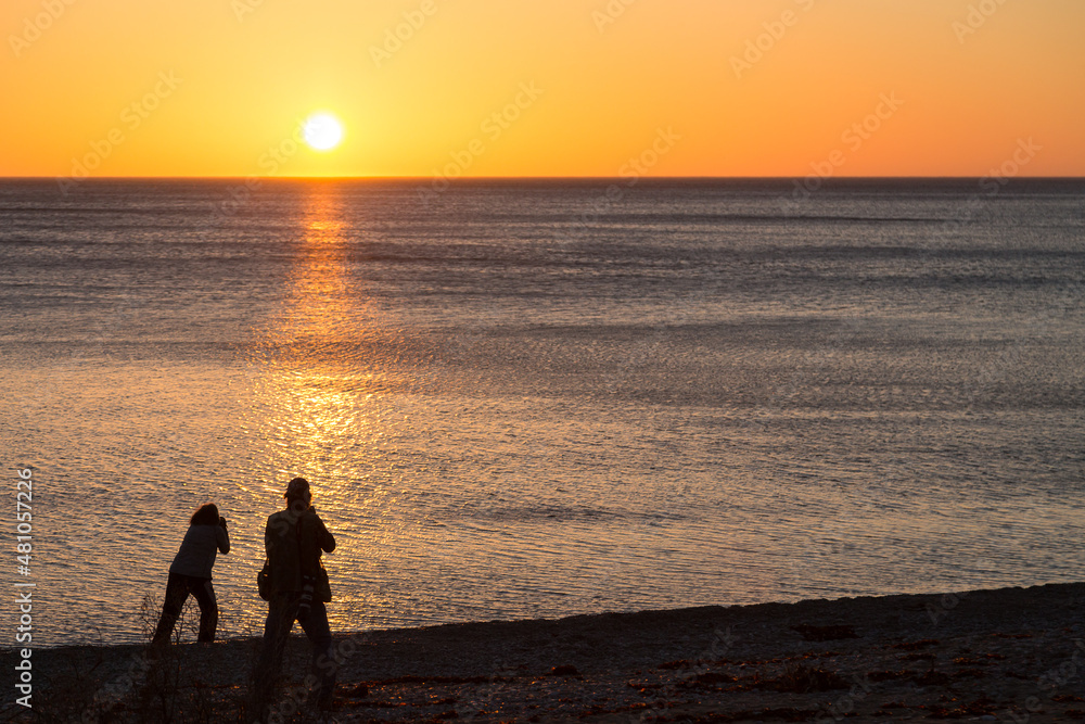 Horizontal back view of man and woman in silhouette taking photos on the St. Lawrence Gulf shore during a beautiful summer sunset, Anticosti, Quebec, Canada