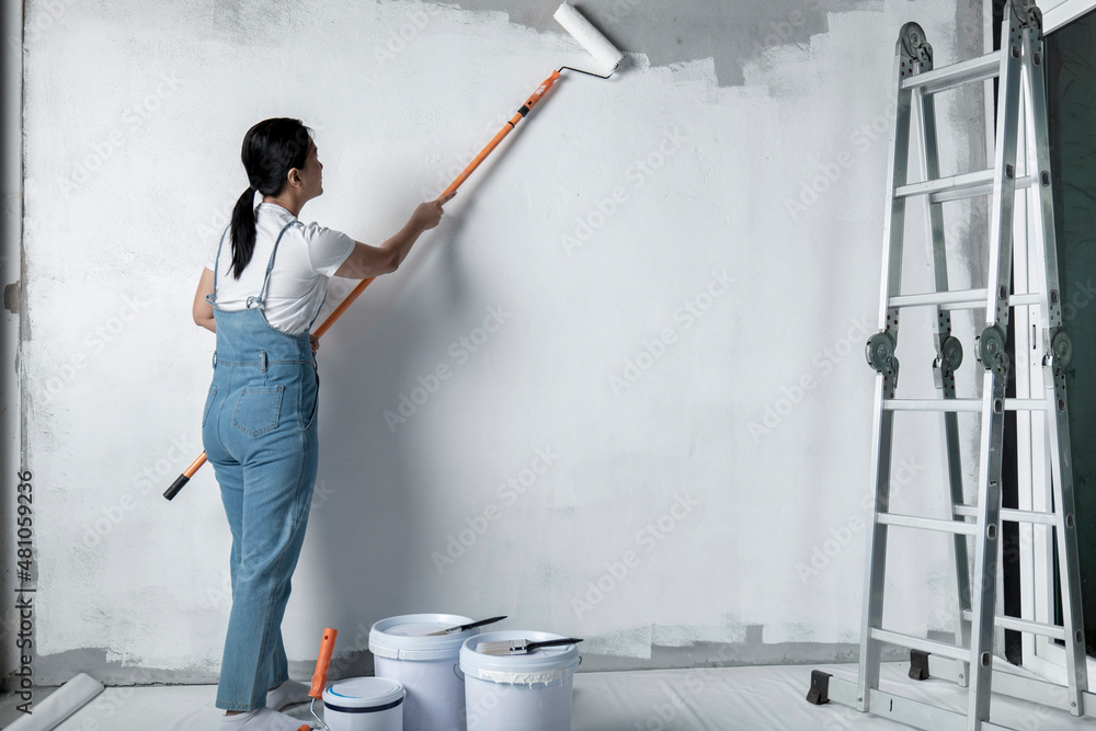 girl paints a white wall with a roller. Repair of the interior. Young  female decorator painting