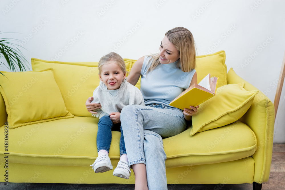 Kid sticking out tongue near mom with book on couch at home.