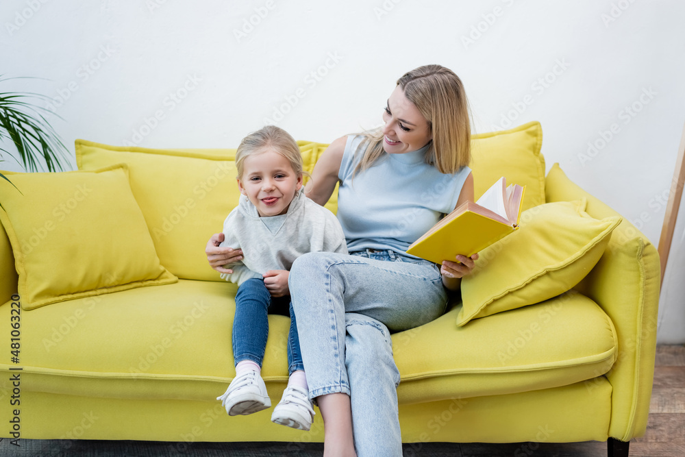 Kid sticking out tongue near mom with book on couch at home. Kid sticking out tongue near mom with book on couch at home.