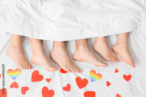 Legs of young man and two women lying in bed covered with paper hearts, top view. Concept of polyamory and LGBT photo