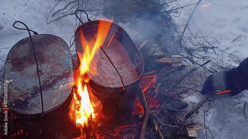 Two cauldrons with camp food on a campfire. Cooking food on fire in a camping trip in nature. Winter hiking and delicious weekend picnic ioutdoors. Gloved hand puts firewood into a bonfire  photo