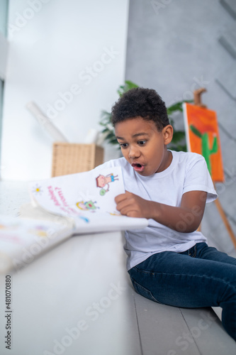 Boy sitting in room looking at book in surprise