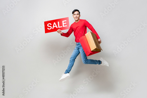 Young Asian man holding shopping bags and red sale sign jumping in isolated light gray studio background for Chinese new year sale concept photo