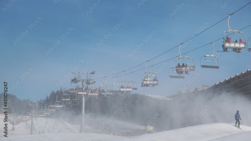 People take ski lift to skiing on mountain slope, sunny winter morning. Background of spruce forest, snow cannons blowing artificial snow of water in frost. Ski resort in white snow. Active vacation People take ski lift to skiing on mountain slope, sunny winter morning. Background of spruce forest, snow cannons blowing artificial snow of water in frost. Ski resort in white snow. Active vacation