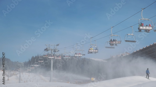 People take ski lift to skiing on mountain slope, sunny winter morning. Background of spruce forest, snow cannons blowing artificial snow of water in frost. Ski resort in white snow. Active vacation