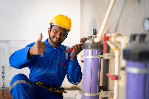 Fotografering smilling young indian engineer or repairman showing thumbs up sign or hand gesture while working at constucation site - concept of professional blue collar job, maintenance service and repair service