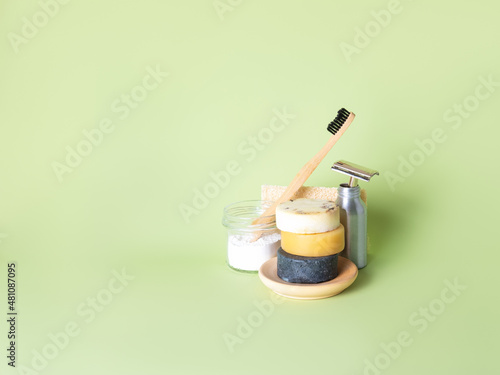 The bamboo toothbrush in glass with toothpowder and steel multiple-use razor in bottle, three natural round soaps and natural wash gourd against the green background. Zero waste bathing concept.
