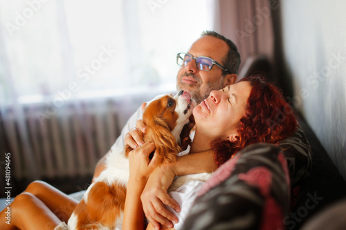 Tableau sur toile cute european middle aged couple in apartment cuddling together with dog cavalier king charles spaniel, cozy time spent by man and woman