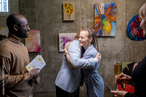 Happy young female embracing modern artist while standing against wall with collection of his paintings in art gallery