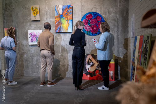 Young blond woman in black casualwear talking to mature elegant man among visitors of contemporary art gallery