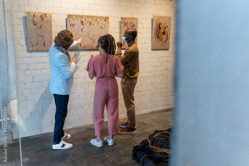 Young intercultural couple in masks listening to mature artist during presentation of his new artwork collection in gallery