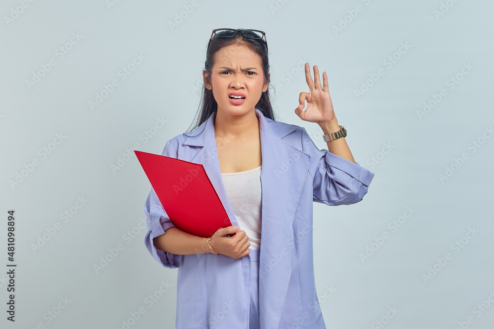 Portrait of confused young Asian business woman in suit holding document folder and makes okay gesture, agrees with nice idea isolated on purple background Portrait of confused young Asian business woman in suit holding document folder and makes okay gesture, agrees with nice idea isolated on purple background