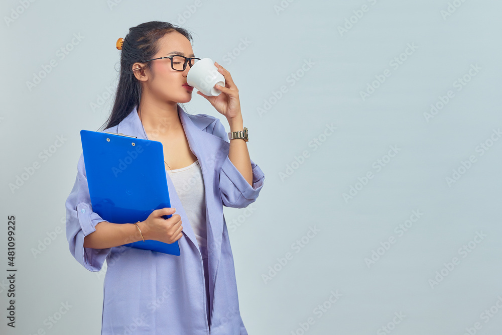 Portrait of beautiful young Asian business woman in suit enjoying cup of hot coffee and holding document folder isolated on purple background Portrait of beautiful young Asian business woman in suit enjoying cup of hot coffee and holding document folder isolated on purple background