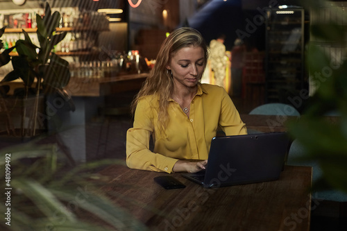 beautiful young caucasian business woman working on laptop. female freelancer in yellow shirt sitting at table, shot through window glass outside of cafe. modern technology, distant work, remote job