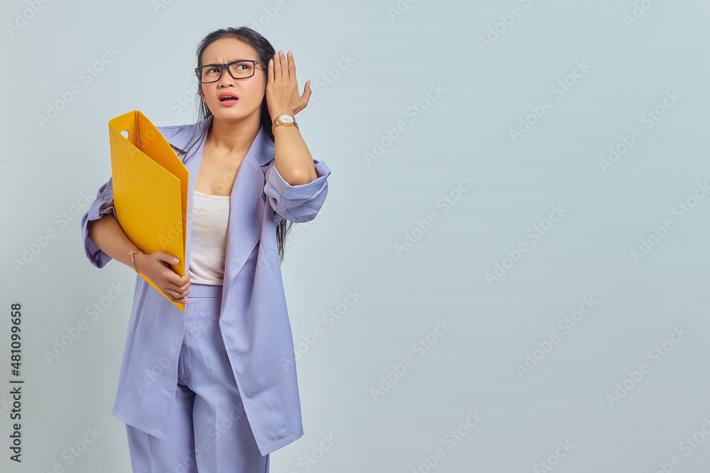 Portrait of serious young Asian business woman keeps hand near ear tries to overhear private conversation listens gossips while holding yellow folder isolated on purple background Portrait of serious young Asian business woman keeps hand near ear tries to overhear private conversation listens gossips while holding yellow folder isolated on purple background